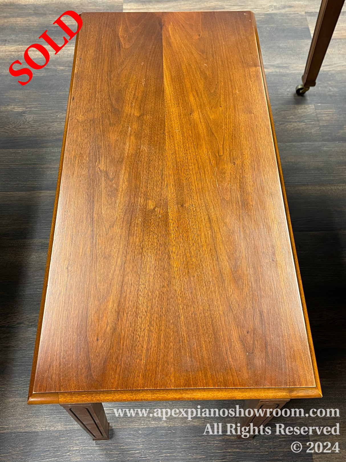 A wooden piano bench with a smooth, polished surface and a rich brown finish, featuring a traditional rectangular design with two visible legs capped with small wheels, indicative of classical styling in piano furniture.