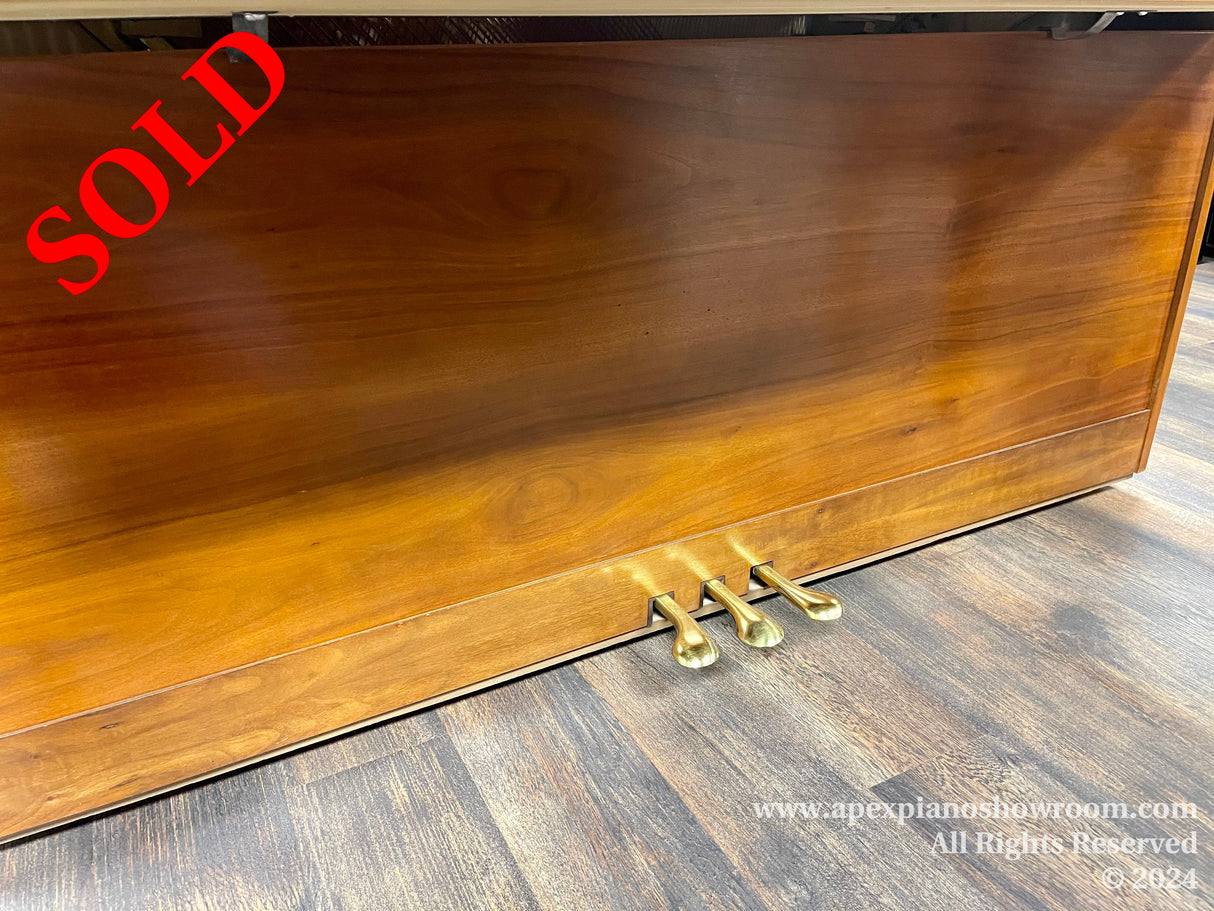 Polished walnut upright piano with brass pedals on a wooden floor, showcasing its sleek finish and elegant design.