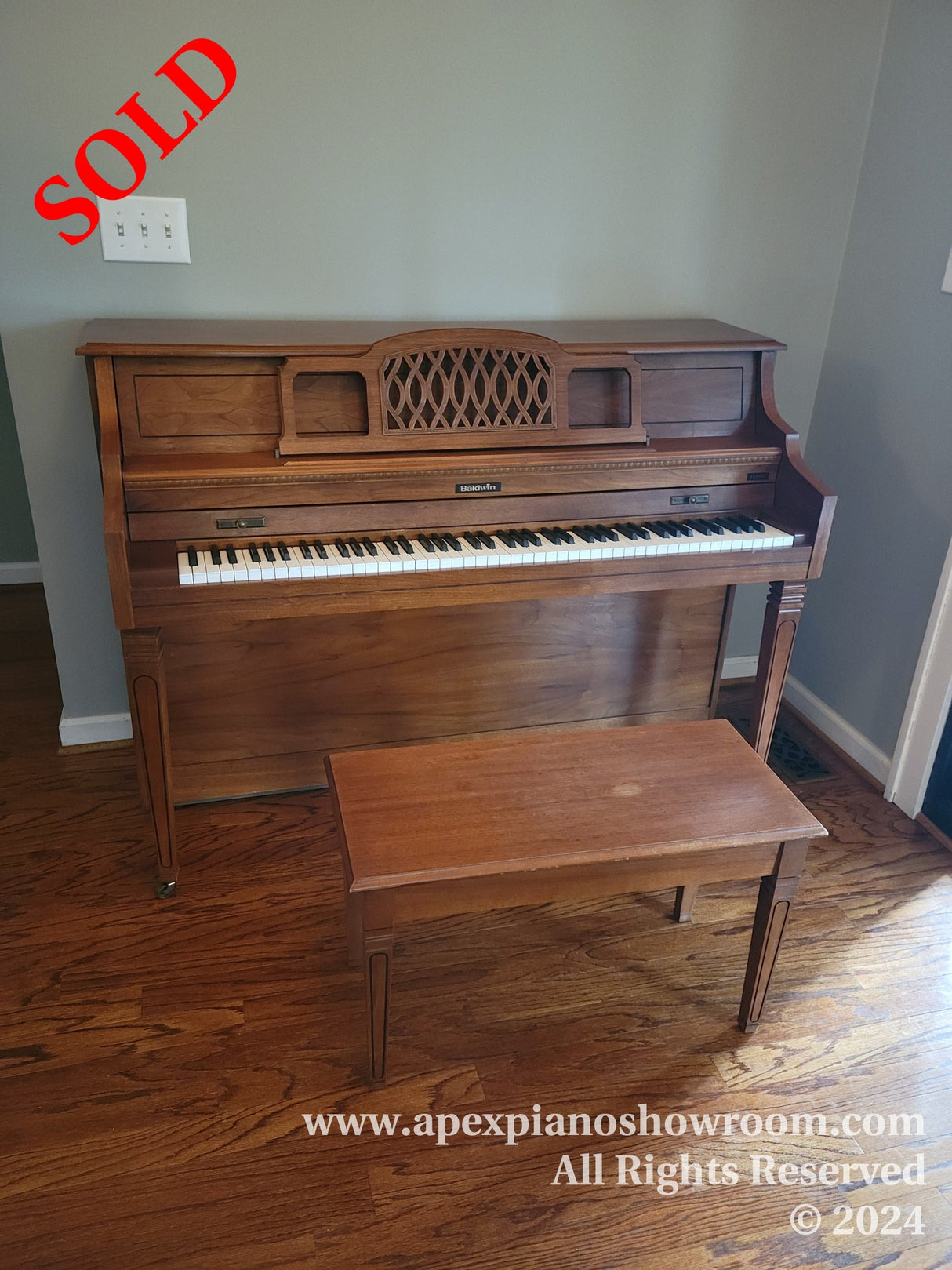 A wooden upright Baldwin piano with decorative lattice music rack, accompanied by a matching wooden bench, placed on a hardwood floor in a room with light-colored walls.