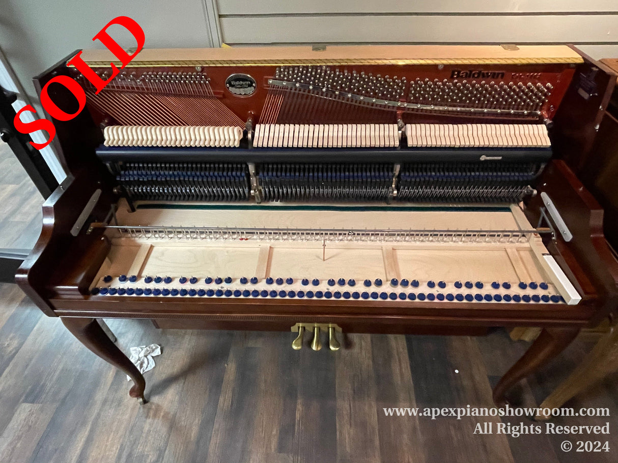 Interior workings of a Baldwin grand piano with its strings, hammers, and dampers exposed, set against a dark wood flooring.
