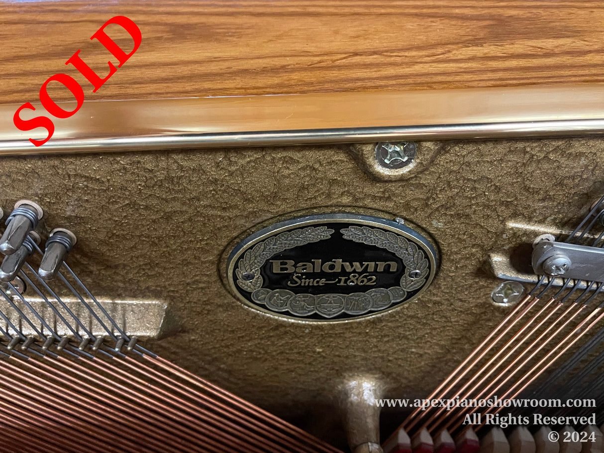 Close-up view of the inside of a Baldwin piano, showcasing the brand emblem with Baldwin - Since 1862 inscription, surrounded by the pianos strings and tuning pins, set against a felt background with the wood finish of the pianos casing visible above.