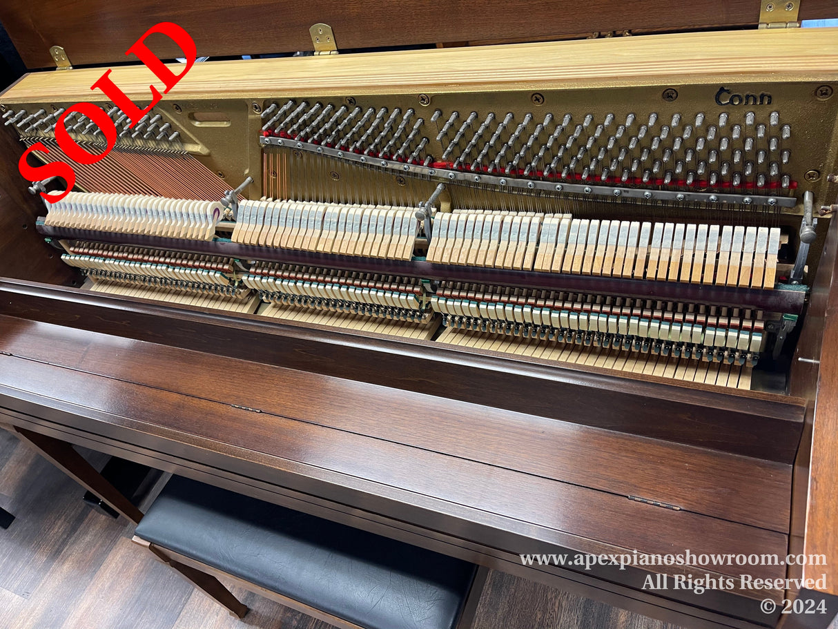 Interior view of an upright piano by Conn showing exposed hammers and strings with intricate details of its internal mechanism, standing on a wooden floor with a matching piano bench underneath.