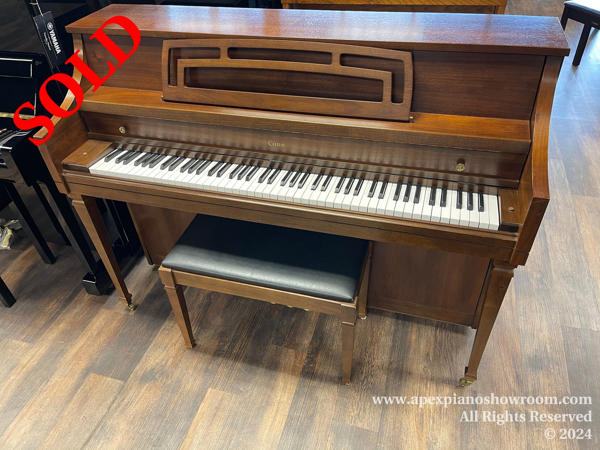 A Conn upright piano with a walnut finish and matching bench positioned on a wooden floor in a showroom setting, showcasing its 88 keys and traditional cabinetry.