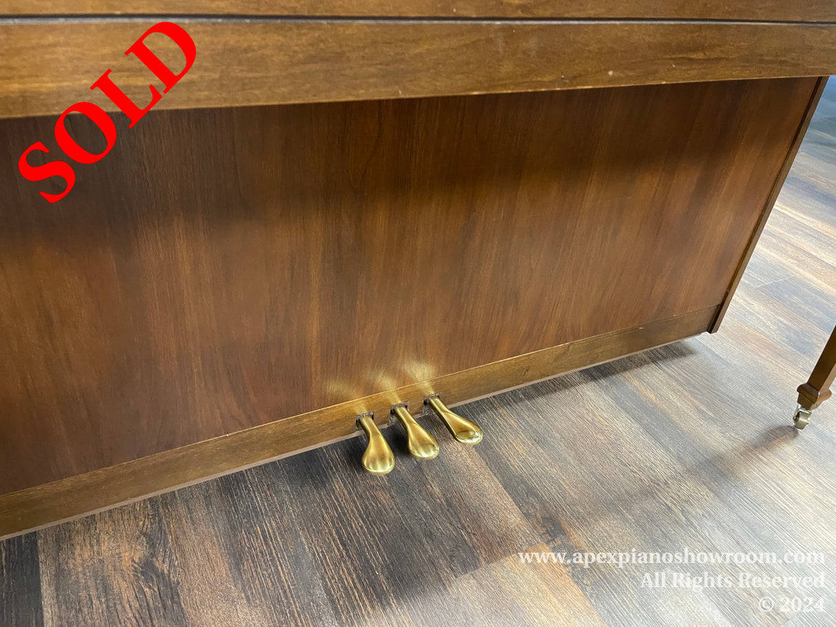 Lower section of a brown upright piano showing three brass pedals and a wooden bottom panel, on a textured gray floor.