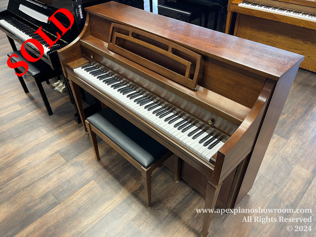 A wooden upright piano with matching bench in a piano showroom, positioned on a wood-patterned floor, flanked by other pianos in the background.
