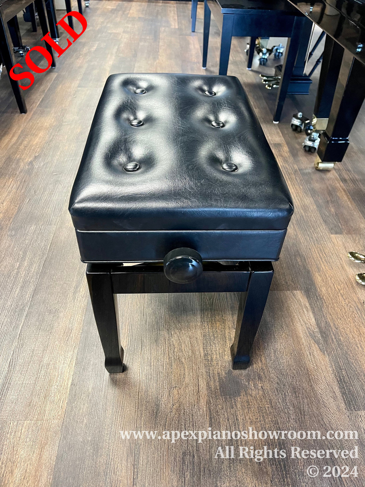 A black, tufted, adjustable piano bench with a storage compartment, positioned in a showroom with wooden flooring next to a polished black piano with caster cups under its legs.