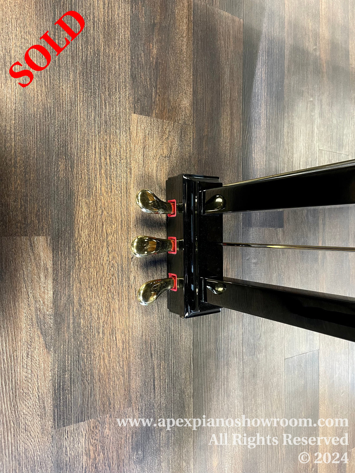 Top-down view of a grand piano with high-gloss black finish and brass casters, positioned on a wooden floor in a showroom, tail end of the piano visible, reflecting overhead lights.