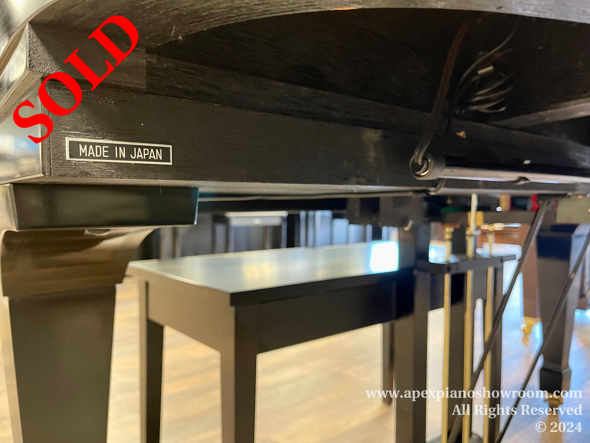 Close-up view of a piano's undercarriage with a Made in Japan label, indicative of the piano's manufacturing origin, set against a blurred background featuring piano legs and a reflection on a polished floor.