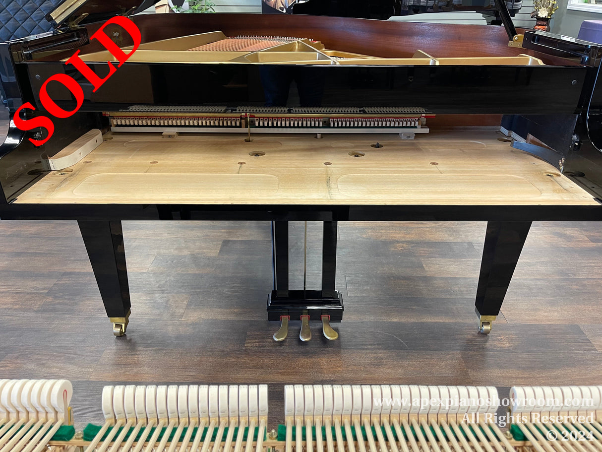 Interior view of a grand piano showing the soundboard, strings, and the action mechanism with pedals visible beneath the keyboard, all set against a dark wooden floor.