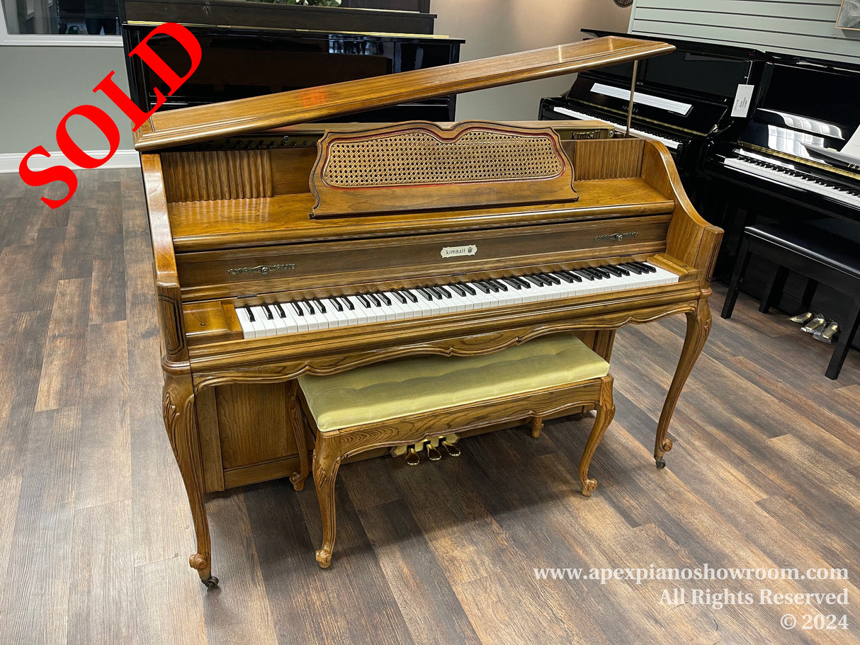 An ornate wooden upright piano with a decorative music desk and curved legs, accompanied by a matching padded bench, displayed in a showroom with other pianos visible in the background.