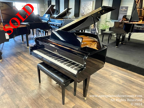 Glossy black grand piano with open lid on display in a piano showroom, surrounded by other pianos, with wood flooring and subtle wall art in the background.