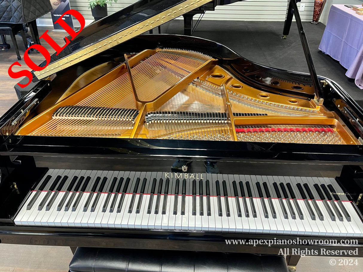 Grand piano with open lid displaying its strings and hammers, alongside a visible brand name KIMBALL on the fallboard, set in a showroom with wood flooring and a table with a purple tablecloth in the background.