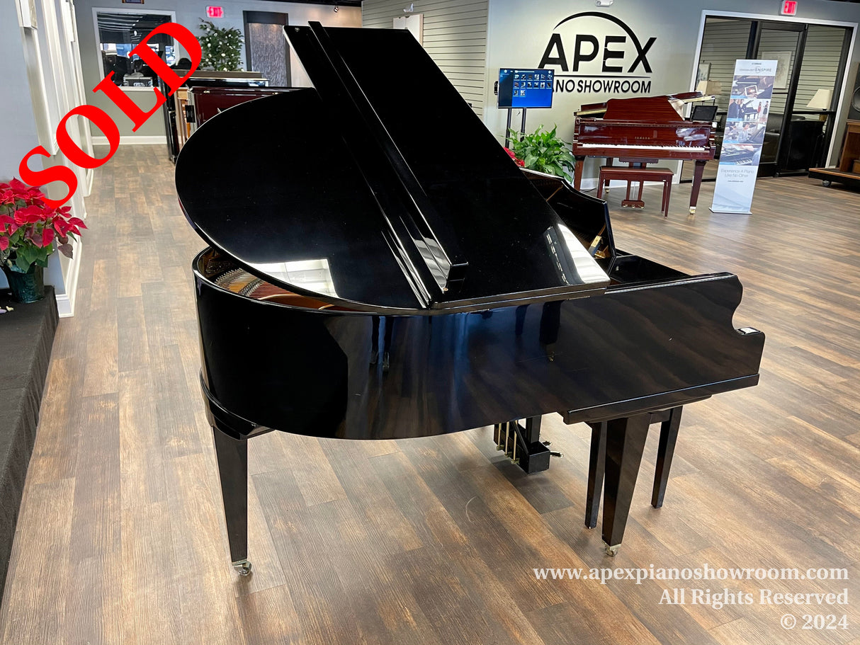 A polished black grand piano prominently displayed in the center of a piano showroom with wood flooring, potted plants, and various pianos in the background, indicating a diverse selection available for customers interested in purchasing a piano.