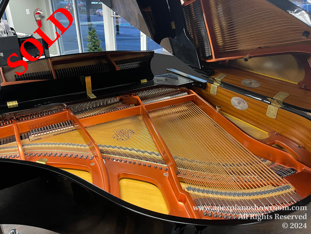 Interior view of a grand piano showing strings, hammers, and the soundboard with a logo, set in a showroom environment, with brass hardware visible on the open lid and keyboard cover.