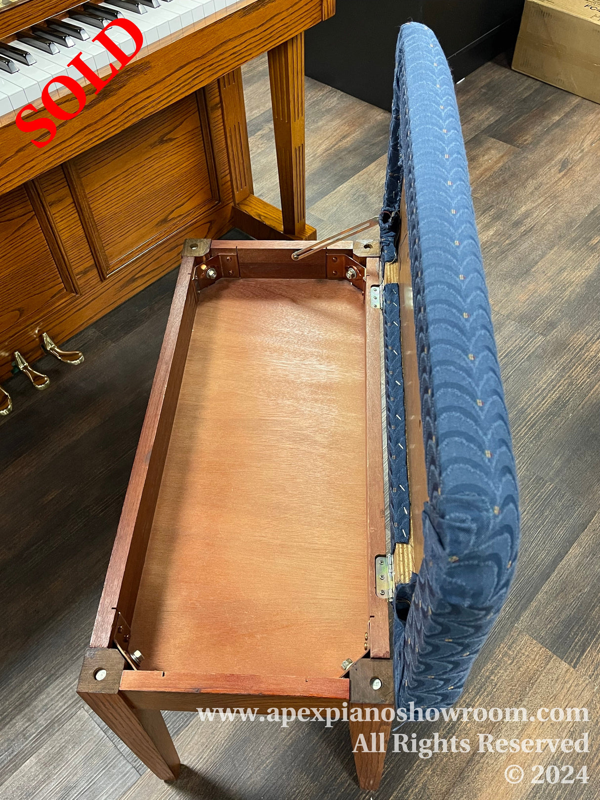 An upright piano with its bench set aside, showcasing the pianos wooden structure and the fabric-covered padded seat of the bench, situated in a piano showroom with hardwood flooring.