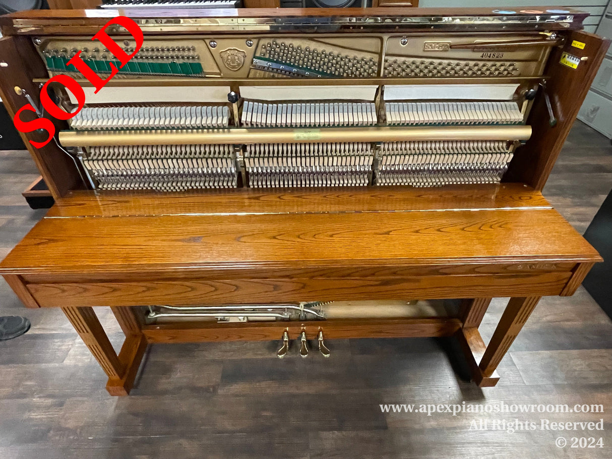 Upright piano with the front panel removed, showing internal strings and hammers, with a polished wooden exterior and pedals visible on the bottom — situated in a room with wooden flooring.