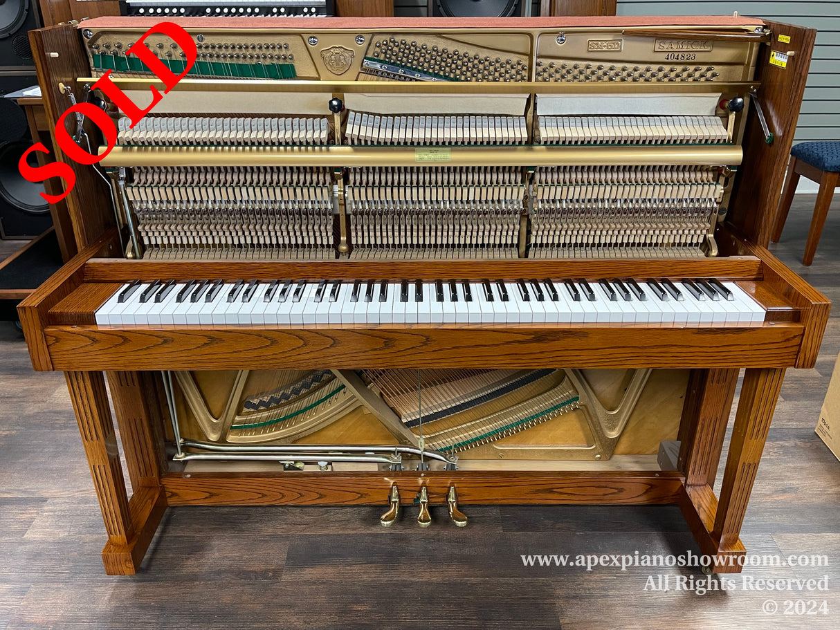 Upright Samick piano with wood finish, white keys visible, internal mechanism exposed, and brass pedal system, set in a showroom environment.