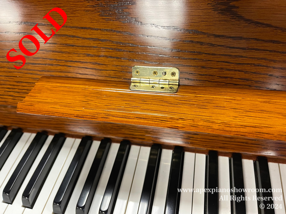 Close-up view of a pianos fallboard with brass hinges, showcasing the wood grain pattern and adjacent black and white piano keys.