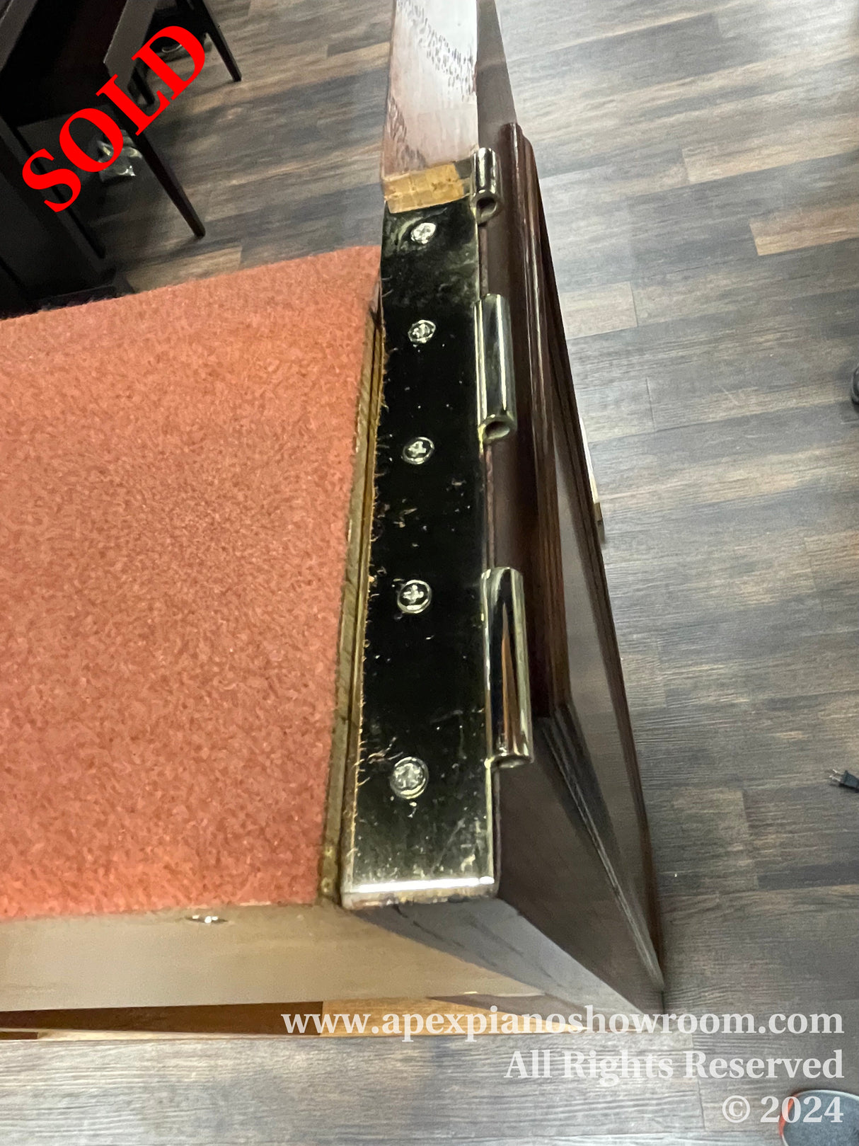 Close-up view of a pianos corner, showcasing the wood finish and metal hardware, with a protective carpet underneath, set against a wooden floor background.