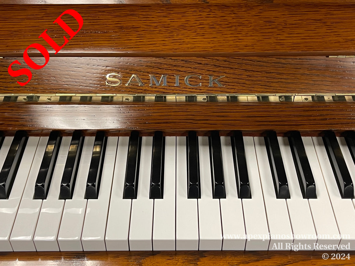 A close-up view of a Samick piano, focusing on the brand name, the polished wooden fallboard, and the black and white keys.