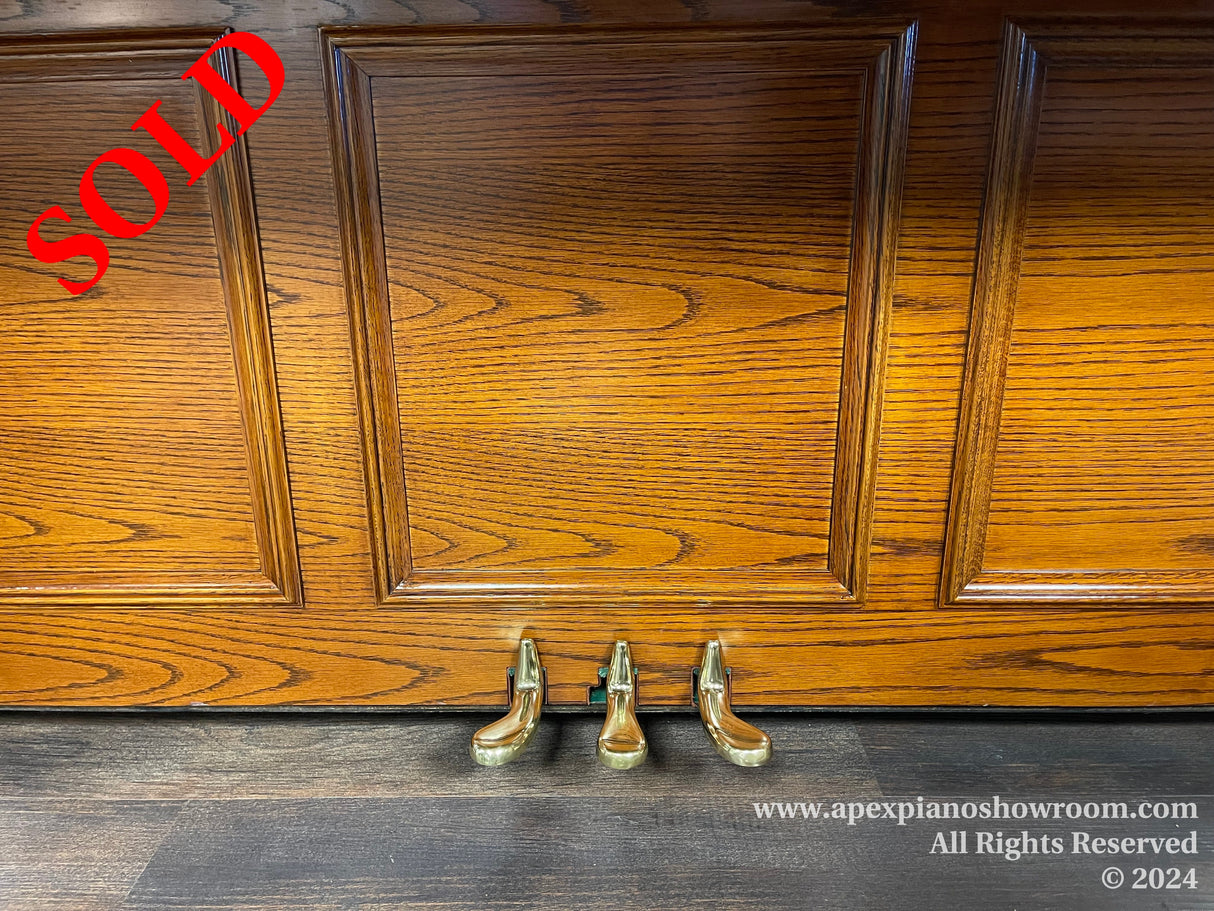 Polished wooden panel of an upright piano with brass pedals showing at the base, set against a hardwood floor in a showroom environment.