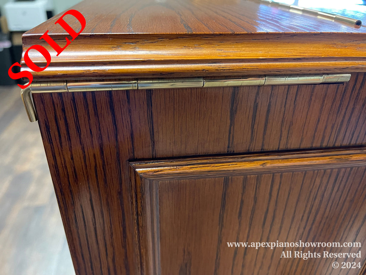 Close-up view of the corner of a wooden upright piano, highlighting the wood grain finish and decorative metal hinges on the panel.