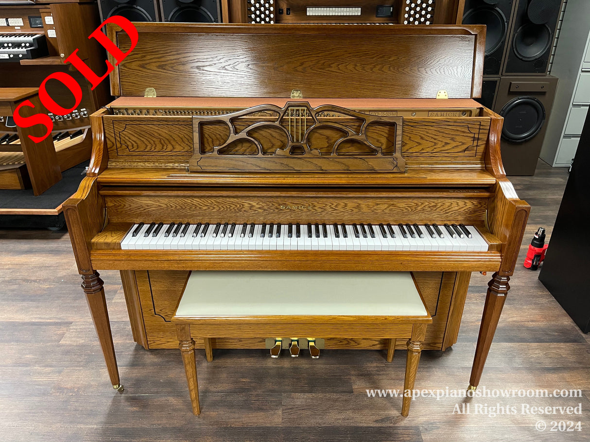 Wooden upright piano with a matching bench in a showroom setting, featuring ornate music desk and carved panel details, set against a backdrop of speakers and electronic equipment.