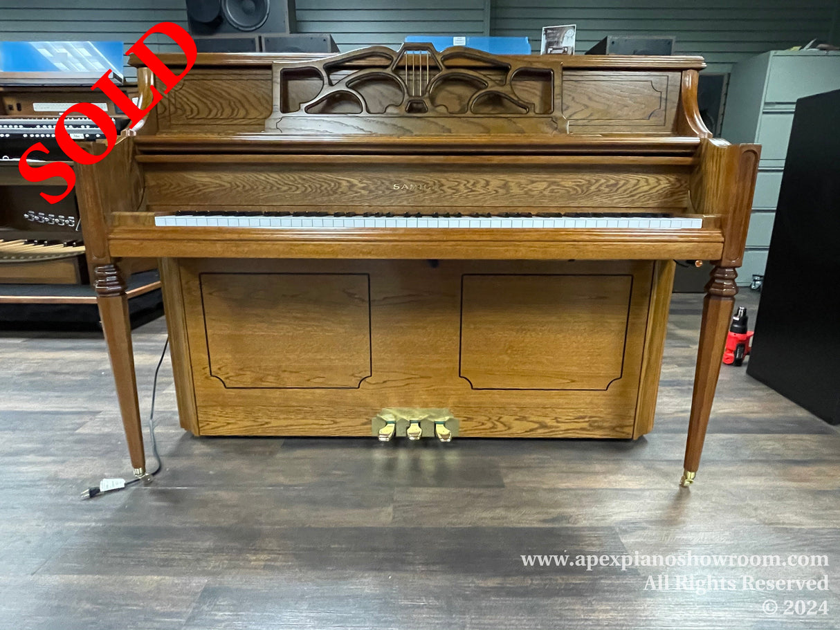 An upright wooden piano with ornate detailing on the music rest, fluted legs, and brass caster wheels, displayed in a showroom setting.