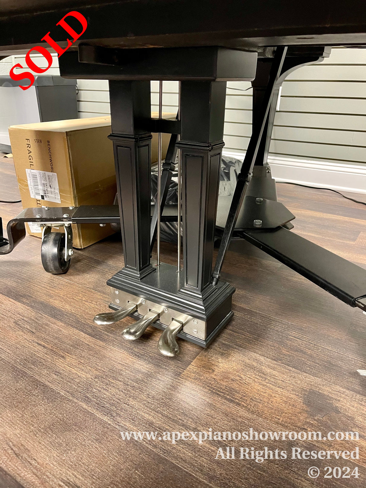 Black grand piano leg with shiny metal casters on a wooden floor, situated in a piano showroom with a cardboard box in the background.