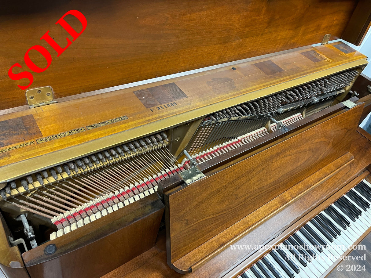 Interior view of a brown upright piano with the front panel removed, revealing the hammers, strings, and inner mechanism — keys are visible at the bottom, and the piano action assembly with felt hammers is prominently displayed.