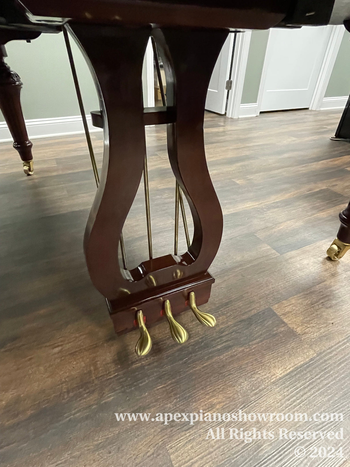 Elegant mahogany grand piano with lyre-shaped pedal assembly and brass casters, showcased on a wood-patterned floor.