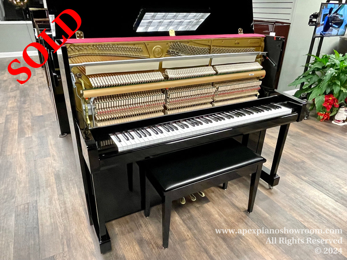 A glossy black upright piano with its internal mechanism exposed, displaying hammers and strings, situated in a showroom with a wooden floor and a potted plant nearby.