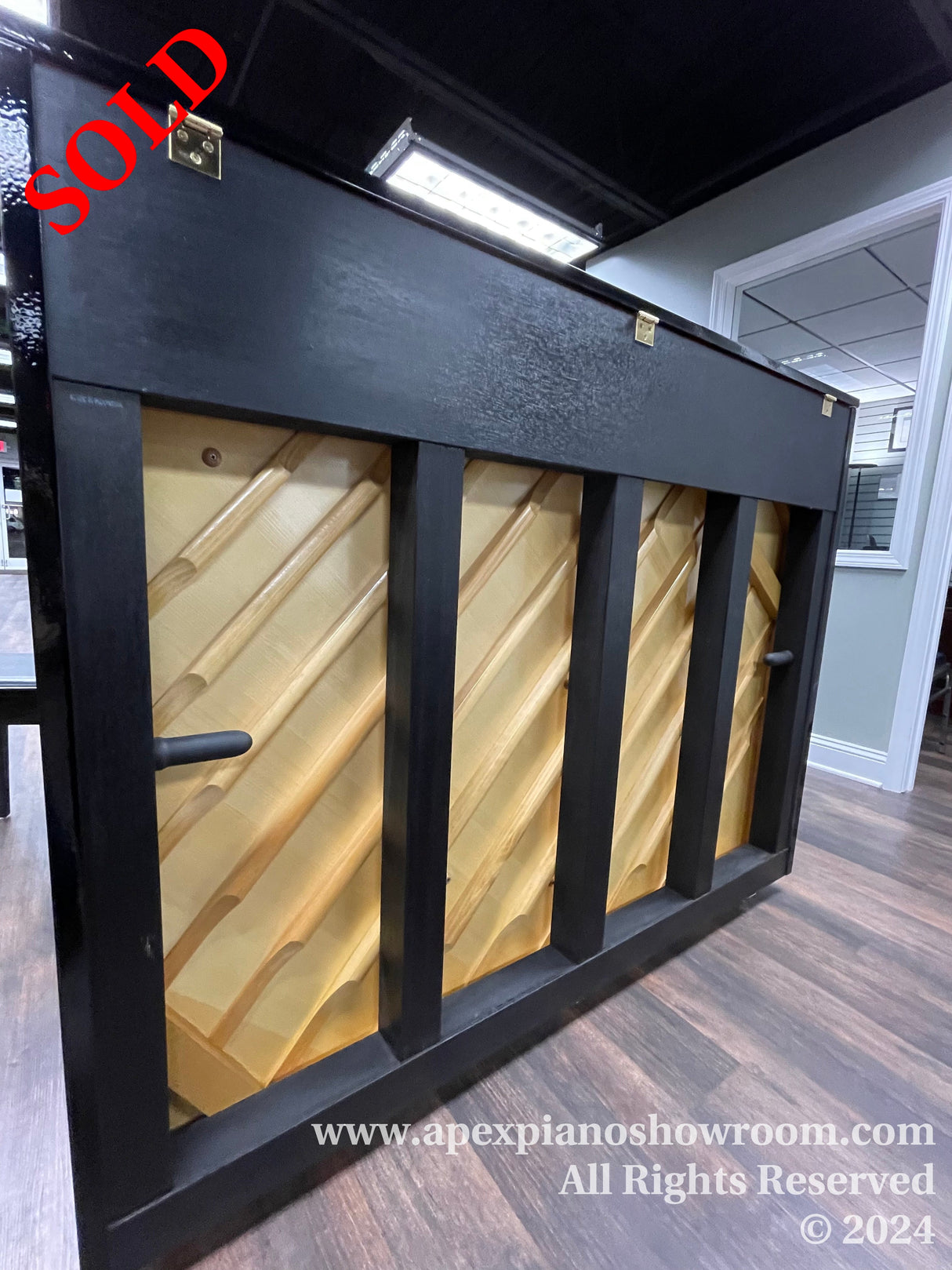 A black upright piano with a golden inner frame on display in a showroom, featuring black vertical bars over the soundboard and under-cabinet lighting, with a company watermark at the bottom.