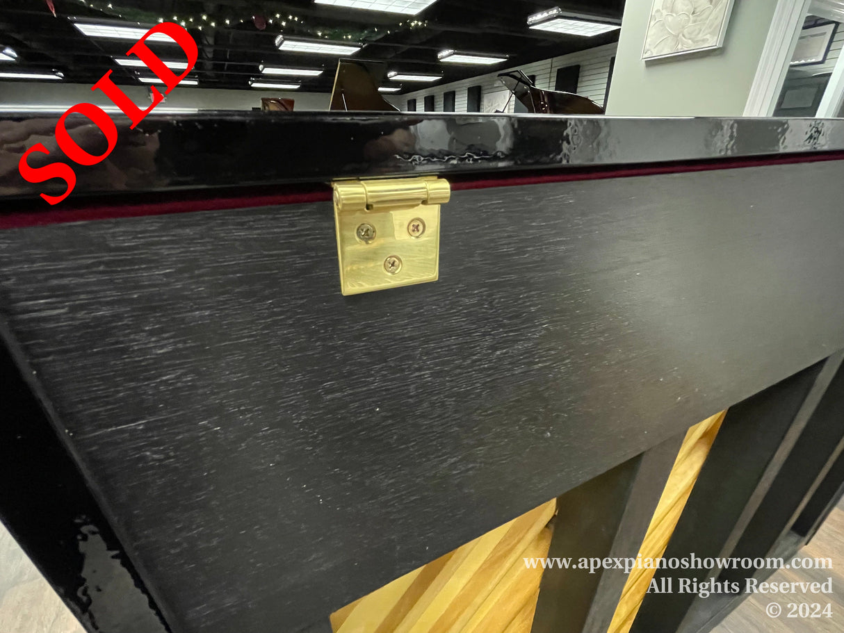 Close-up of a piano fallboard lock with a gold finish on a black piano with subtle wood grain texture, highlighting the locking mechanism used for securing the keyboard.