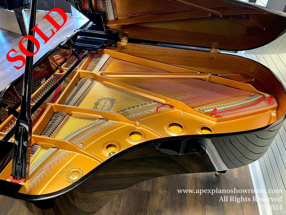 Grand piano interior view showing gold-painted frame, soundboard, strings, and high-tension tuning pins, displayed in a showroom setting.