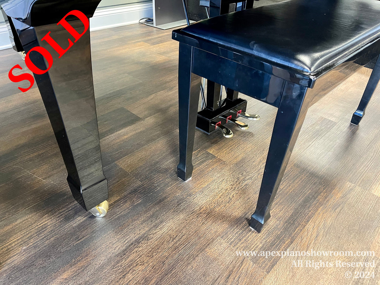 A high-gloss black piano bench with straight legs and brass casters on a wooden floor, positioned next to a piano with pedals visible in the background.