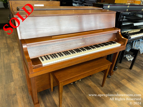 A Yamaha upright piano with a wooden finish displayed in a piano showroom, complete with a matching bench, both set on a dark wooden floor, with various pianos in the background.