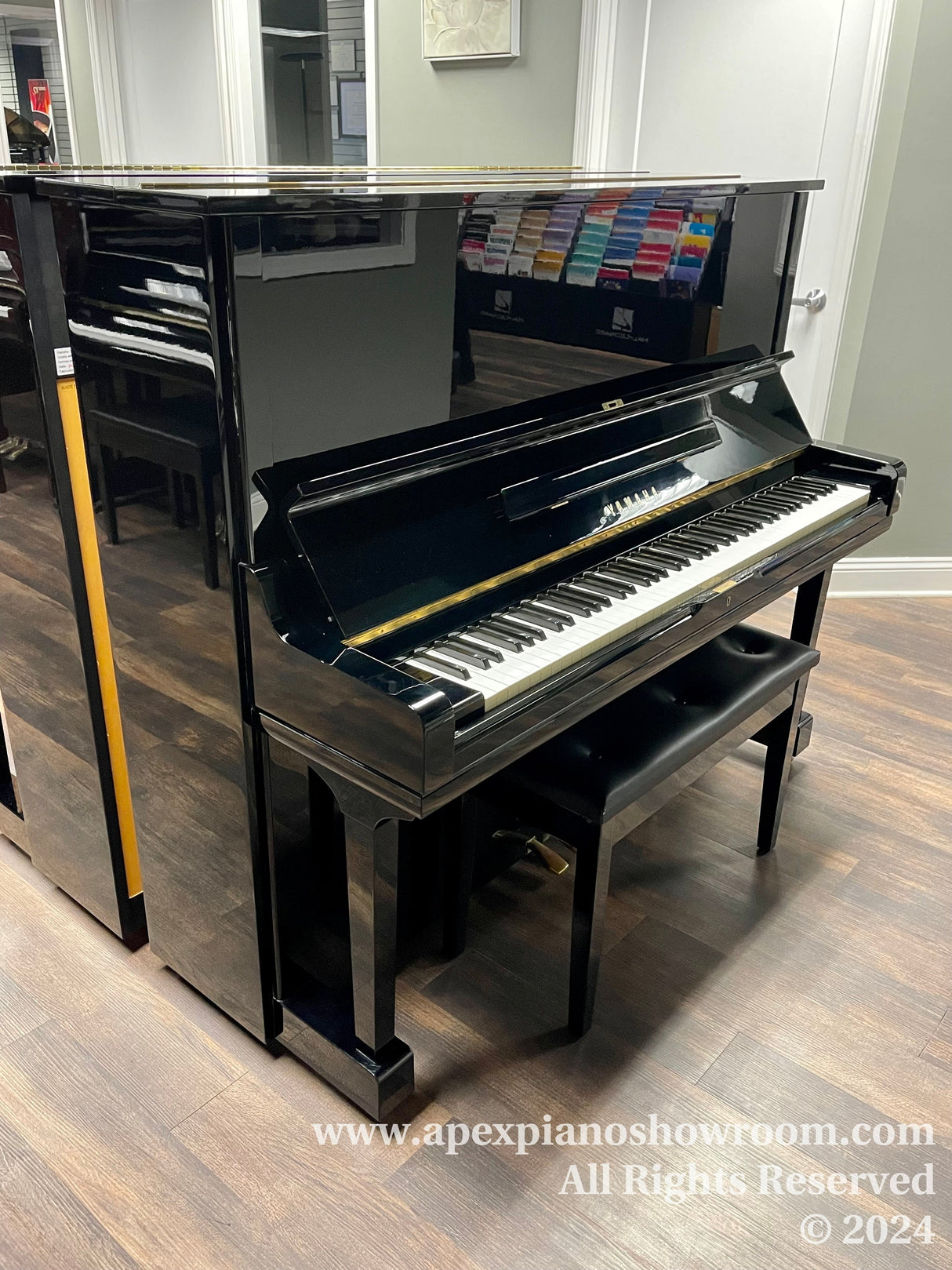 A glossy black upright piano with an open lid revealing the keyboard, set against a showroom backdrop with music books in the background, positioned on a wooden floor.