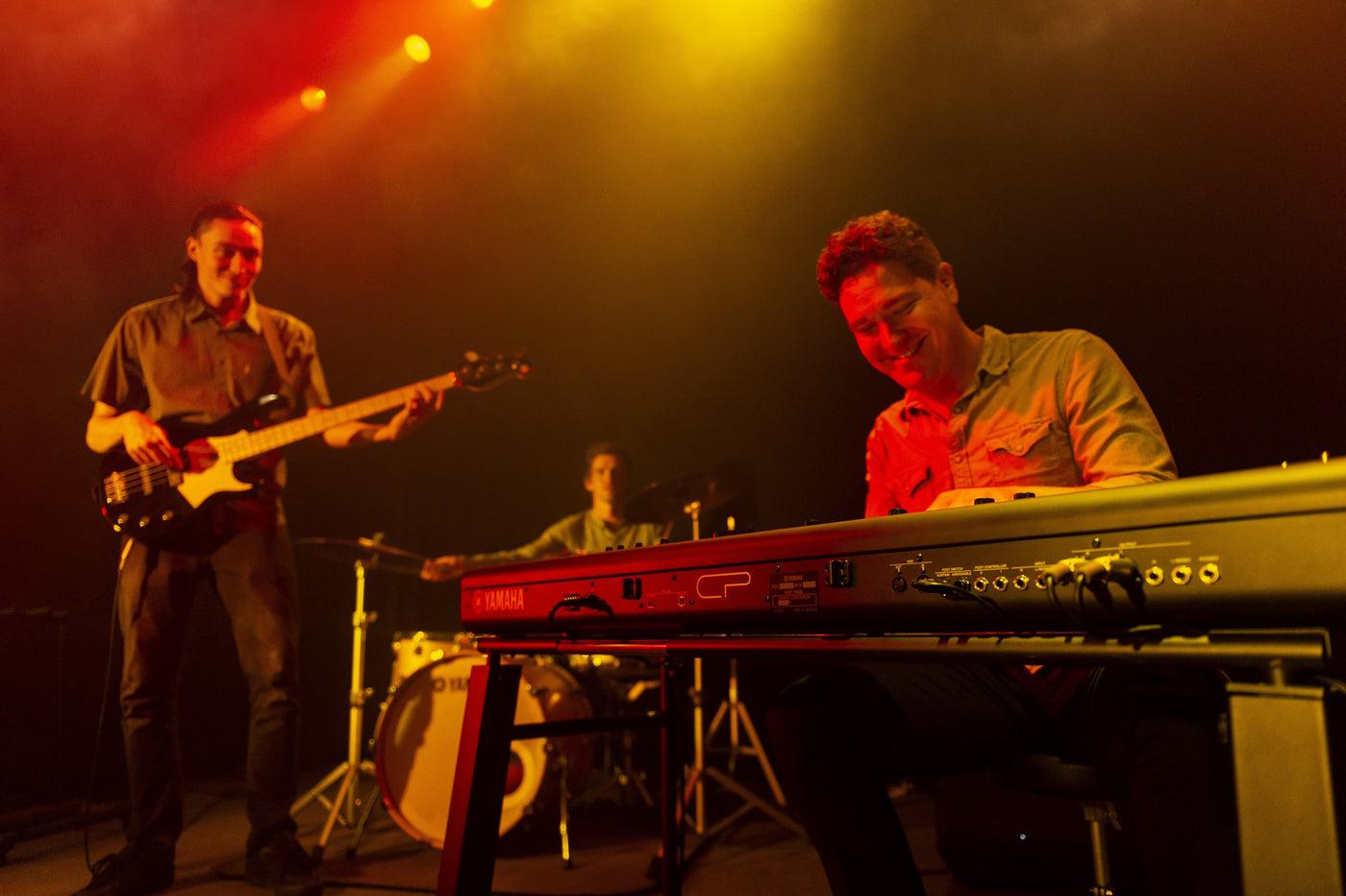 Electronic stage piano being played by a musician on stage with warm lighting, with other band members in the background including a bass guitarist and a drummer.