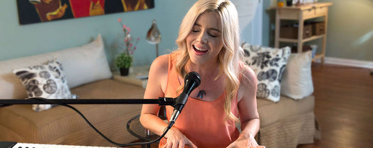 Woman singing into a microphone with a keyboard in front of her, in a cozy living room setting.