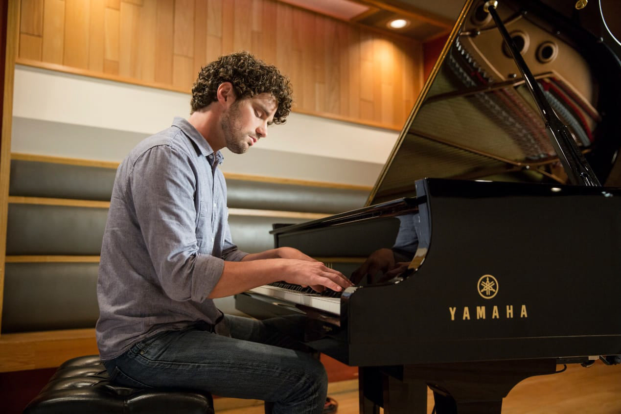 Man playing a Yamaha grand piano in a room with wooden floors and sound panels.