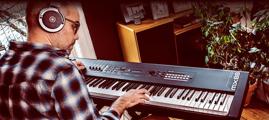 Man playing an electronic keyboard with headphones in a home setting surrounded by houseplants and framed pictures.
