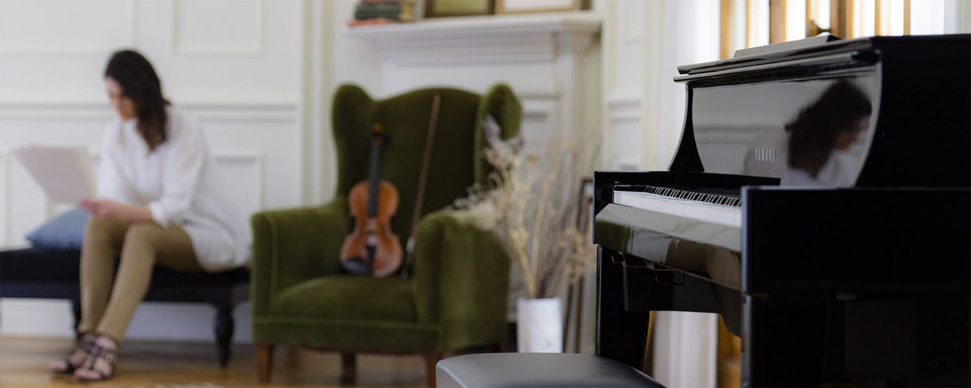 A spacious room featuring a Yamaha grand piano in the foreground with a violin placed on a green armchair, and a blurred figure of a woman reading music sheets in the background, suggesting a setting for a music practice session.