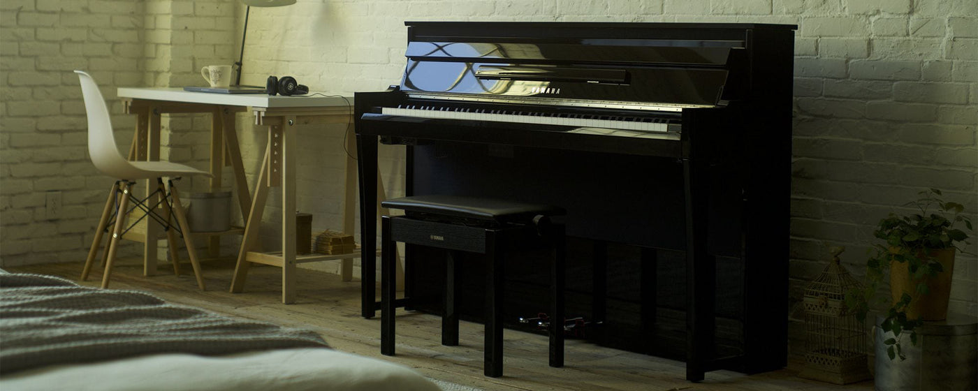 A modern upright piano positioned in a cozy home interior with minimalist furnishings and a white brick wall, ideal for a personal practice space.