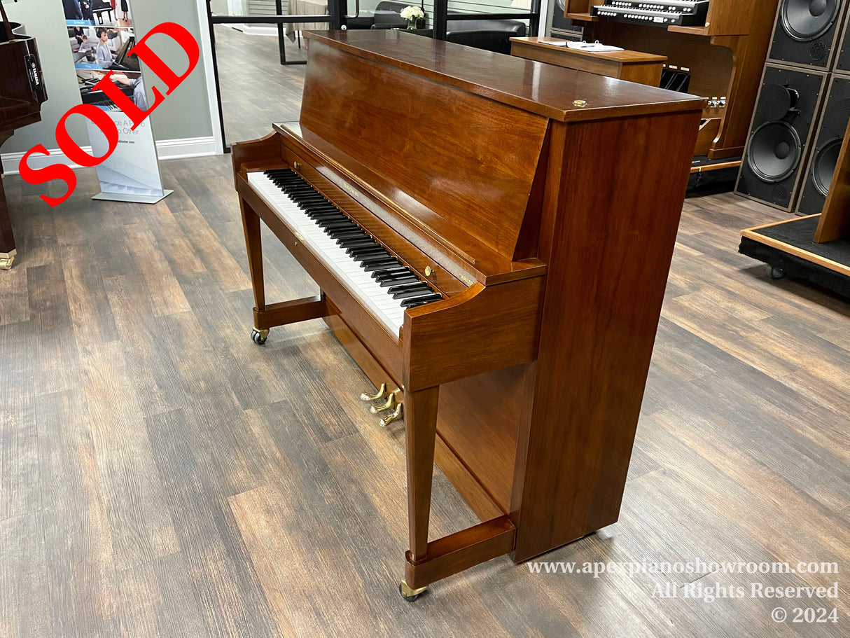 A polished mahogany upright piano with brass fittings on display in a piano showroom, featuring a full keyboard and a raised top lid revealing the internal strings and hammers.
