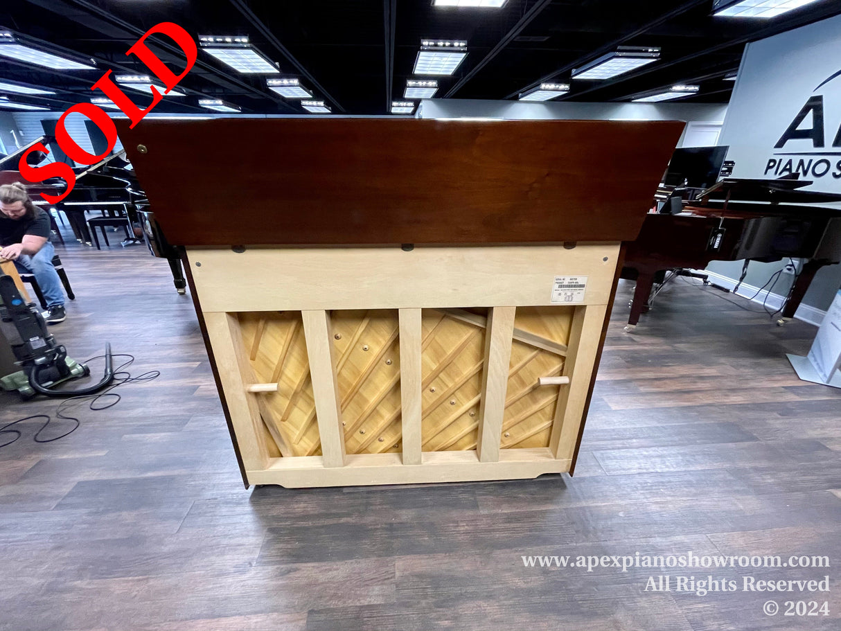 A wooden upright piano displayed in a showroom with a hinged top open, revealing the inside of the piano, set against a backdrop of other pianos and a man seated nearby.