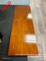 A glossy finished piano fallboard with rich mahogany color, resting on the floor against a black mat and grey wood-patterned flooring.