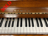 A close-up view of a Samick piano, focusing on the brand name, the polished wooden fallboard, and the black and white keys.