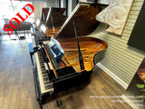 A grand piano with an open lid displaying its shiny black exterior and golden interior, set against a wood-paneled showroom floor with subtle lighting reflecting off the polished surfaces.