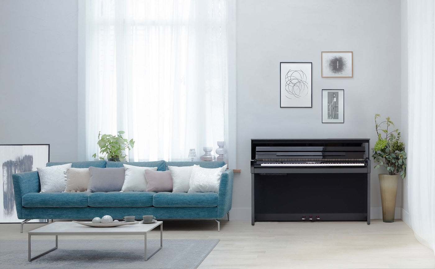 Modern living room featuring a Yamaha Clavinova CLP-785 digital piano in polished ebony. The room has a teal velvet sofa with pastel pillows, a large window with sheer curtains, and minimalist artwork on white walls. The space is decorated with plants and a sleek coffee table, creating a bright, airy atmosphere that balances the piano's presence.